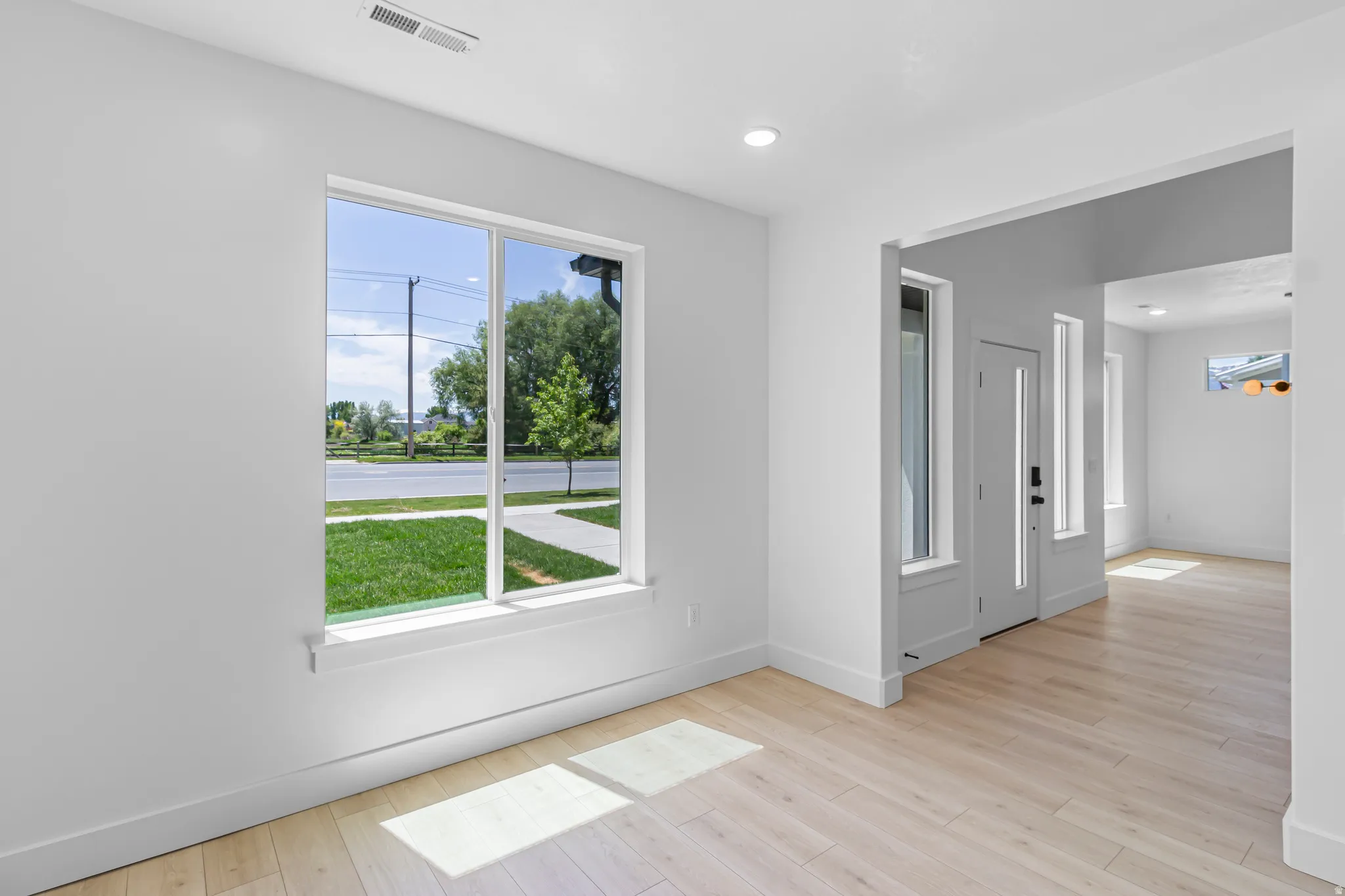 Bright interior space featuring a large window, light wood-finish flooring, recessed lighting, white walls, and a modern entryway with a vertical paneled door and sidelights