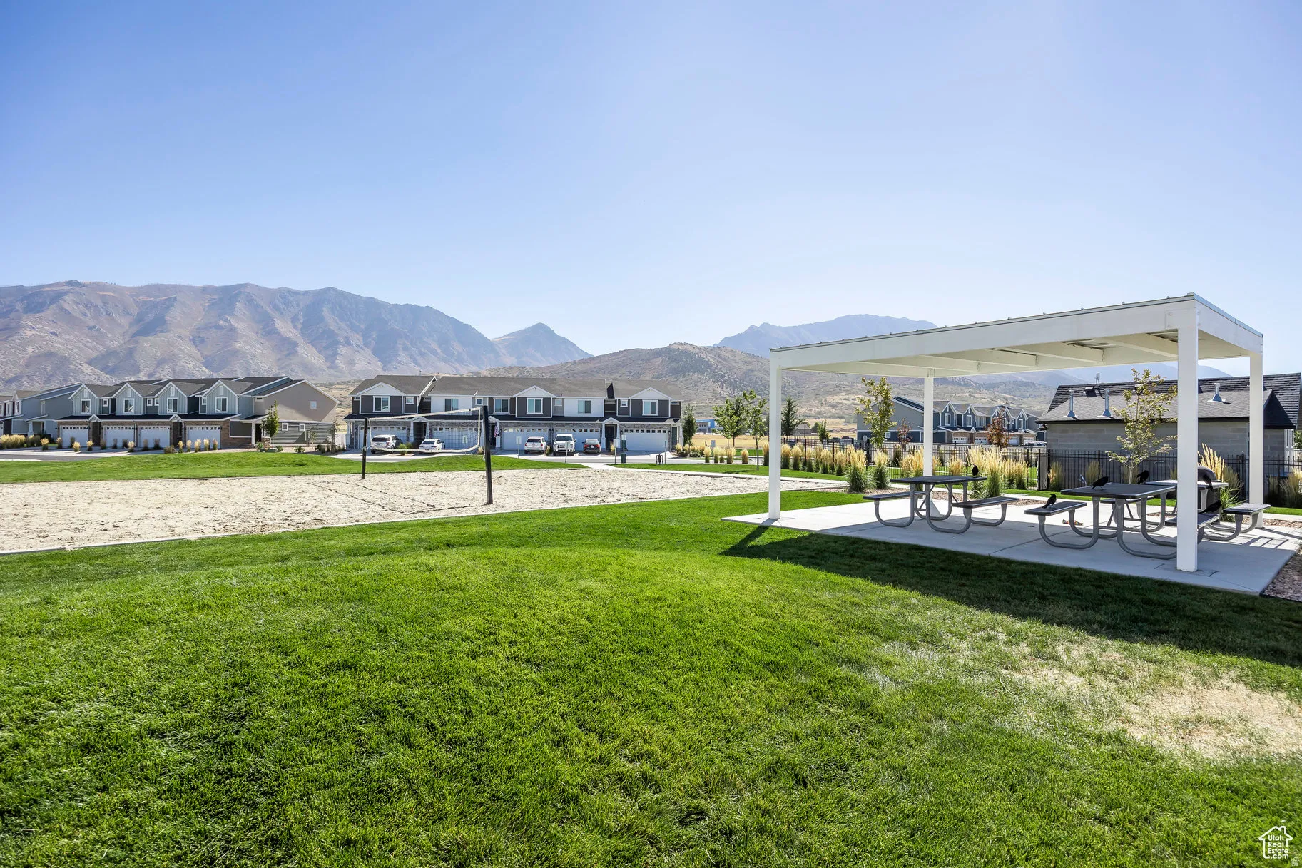 Expansive green space featuring a sand volleyball court, a covered patio area with picnic tables, and distant mountain backdrops