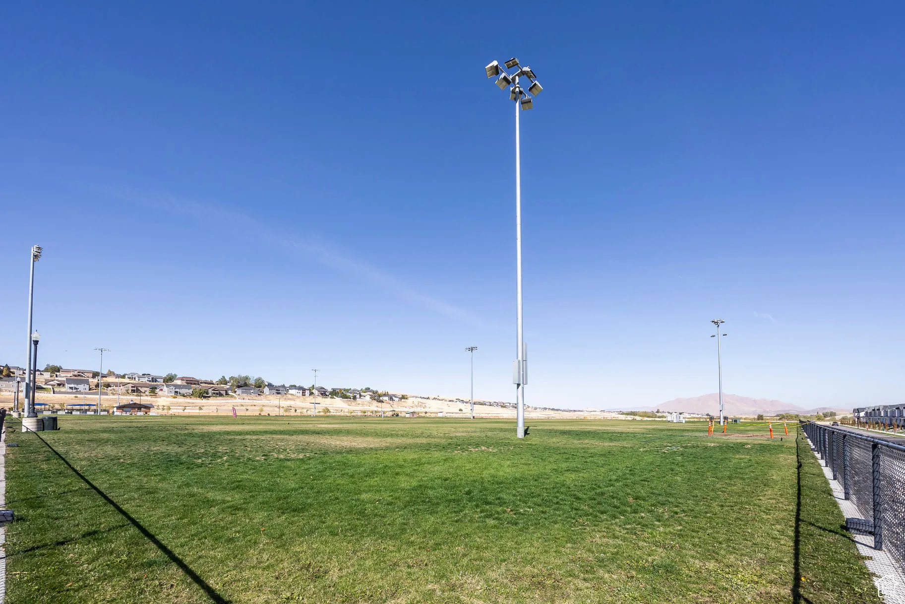 Expansive outdoor field featuring green grass, multiple light poles, a chain-link fence, and distant residential structures