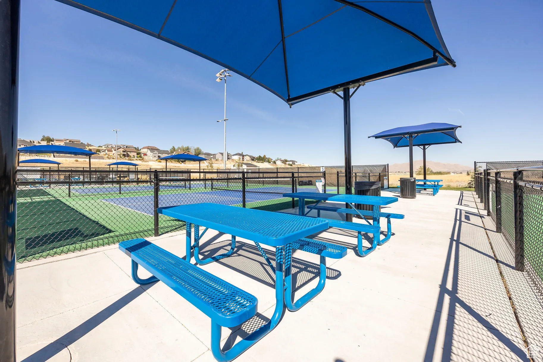 Outdoor recreation area featuring multiple blue picnic tables with benches, overhead shade structures, and concrete paving