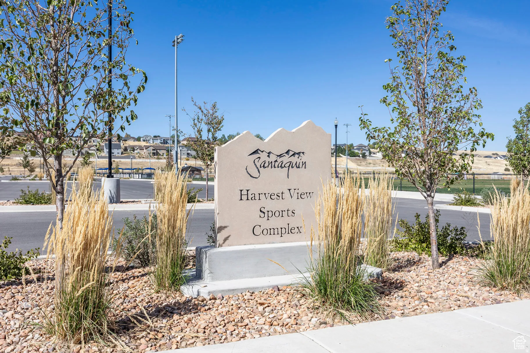 Stone monument sign with sculpted mountain peaks featuring the "Santaquin Harvest View Sports Complex." The sign is set amidst a landscape of low-maintenance gravel, ornamental grasses, and young trees