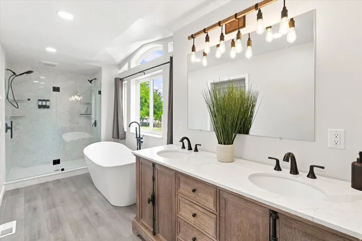 Spacious bathroom featuring a double vanity with stone countertops, a freestanding soaking tub, a glass-enclosed shower with a rainfall showerhead, and wood-finish flooring