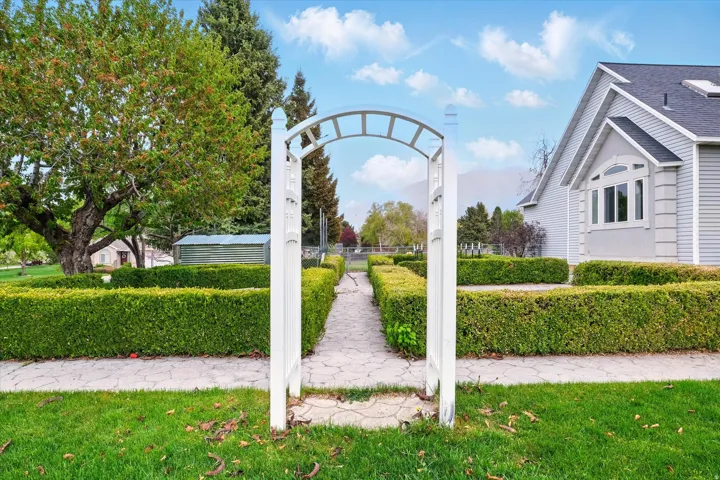 Front yard featuring a white garden archway, patterned concrete walkway, manicured hedges, and mature trees