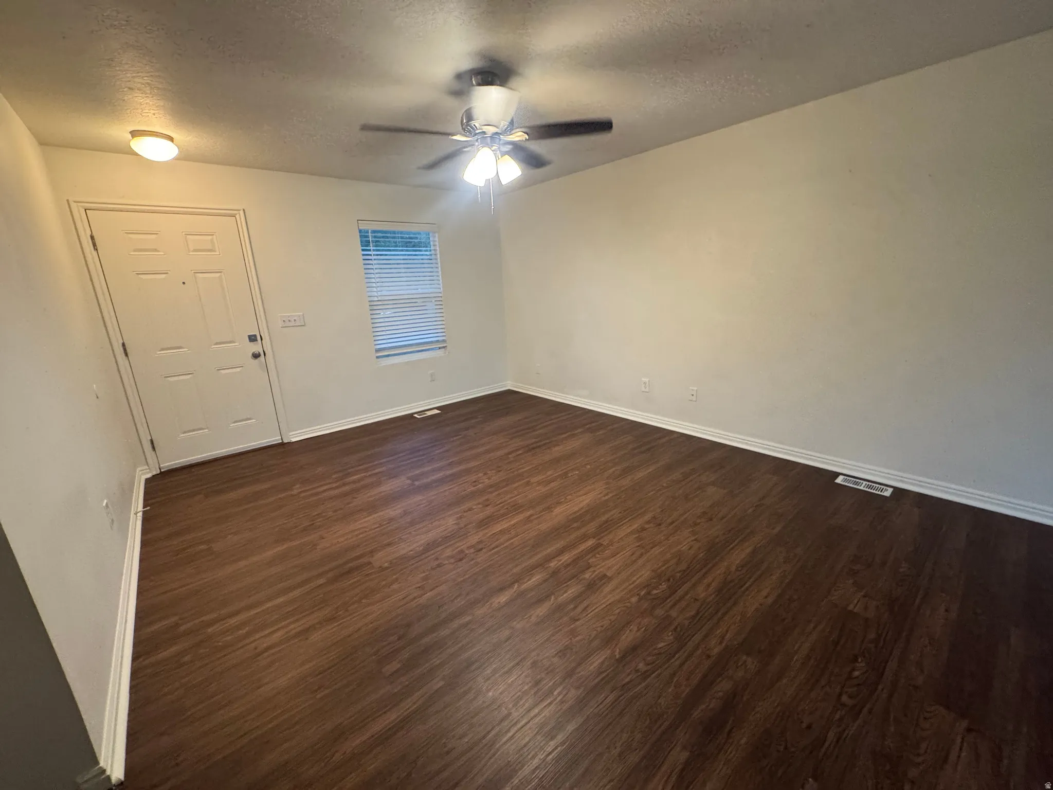 Spacious room featuring rich wood-finish flooring, a ceiling fan with integrated lighting, a white panel door, and a window with blinds