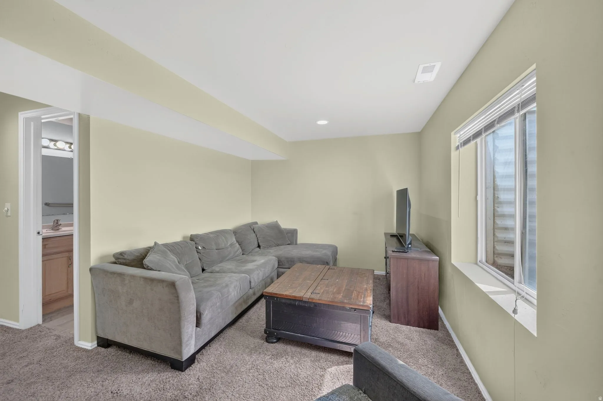 Carpeted living area featuring light-toned walls, recessed lighting, and a single window with blinds