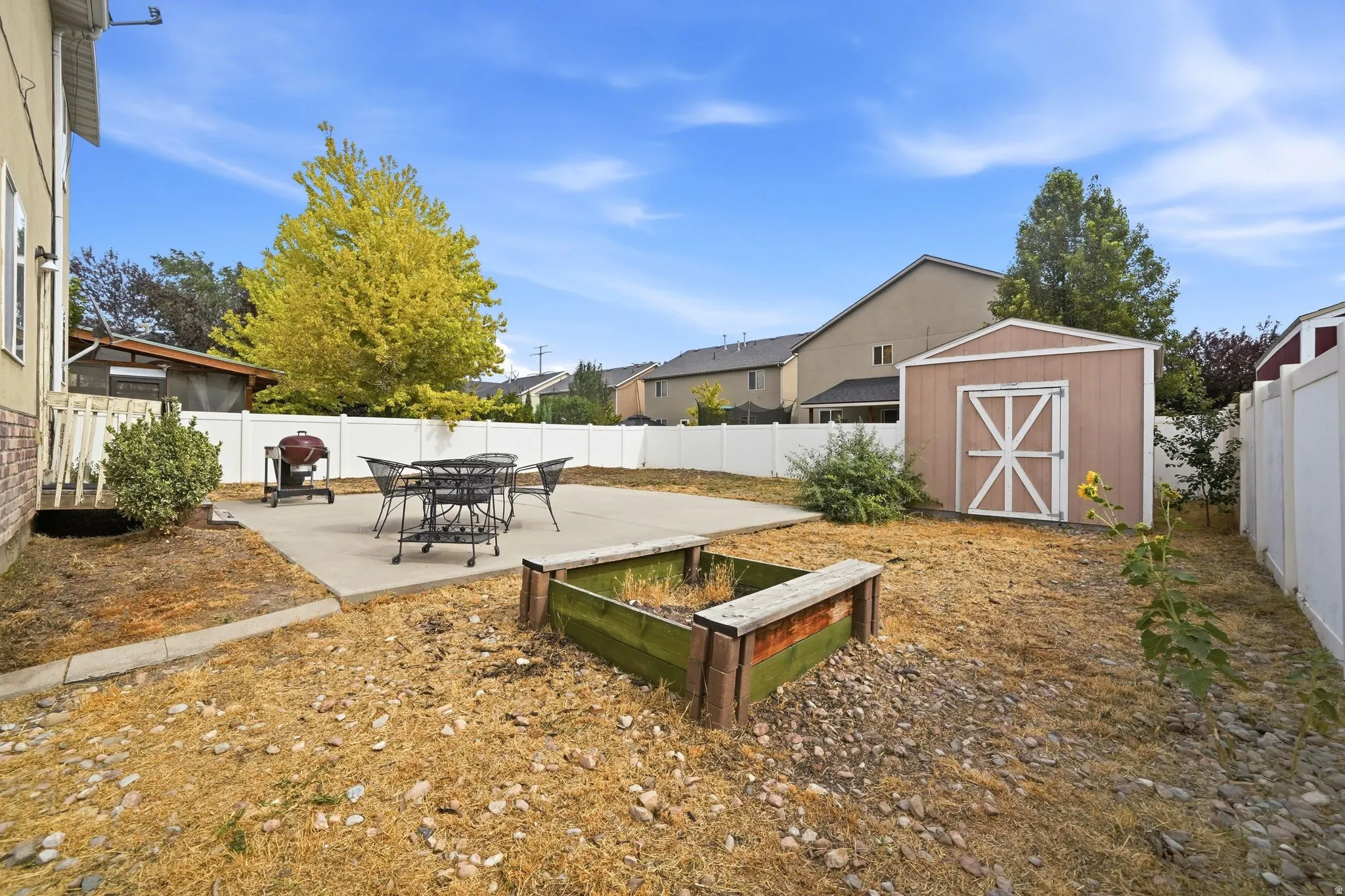 Spacious backyard oasis featuring a concrete patio, sandbox with bench seating, dining table with umbrella, and mature landscaping