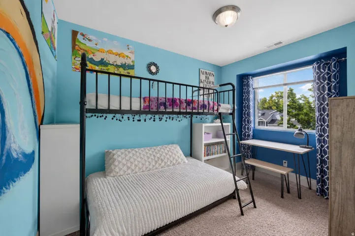Bedroom featuring a black metal bunk bed, light-toned carpet flooring, and a large window with patterned drapes