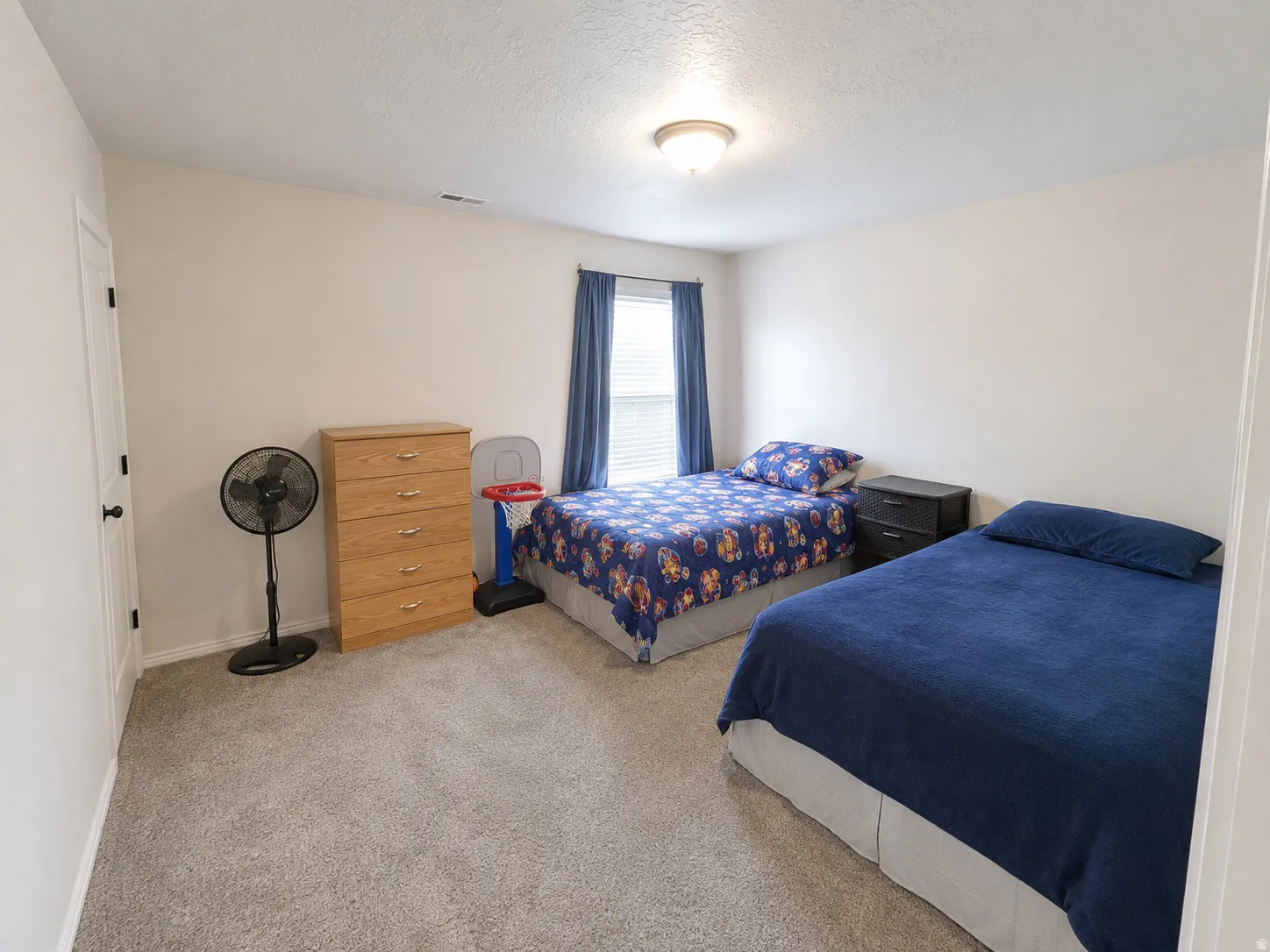 Spacious bedroom featuring carpeted flooring, dual beds with blue bedding, wooden dresser with ample storage, pedestal fan, and large window with blue curtains providing natural light
