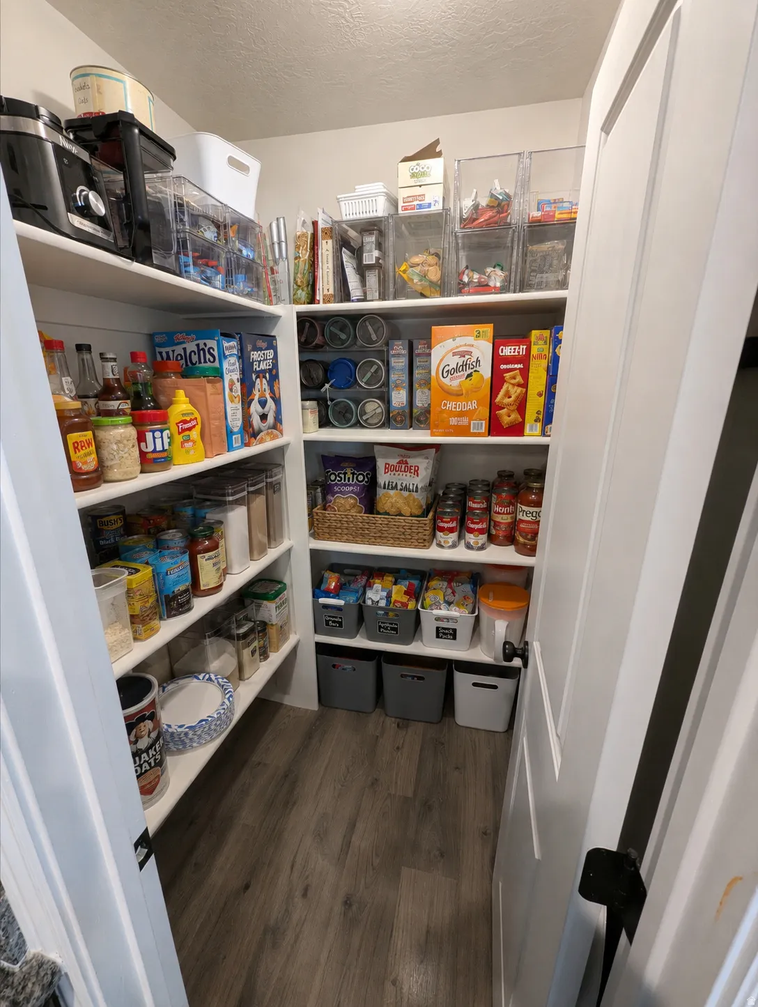 Spacious pantry featuring floor-to-ceiling shelving with multiple storage tiers, built-in cabinetry with closed storage boxes, wood-finish flooring, and ample room for pantry staples and kitchen supplies