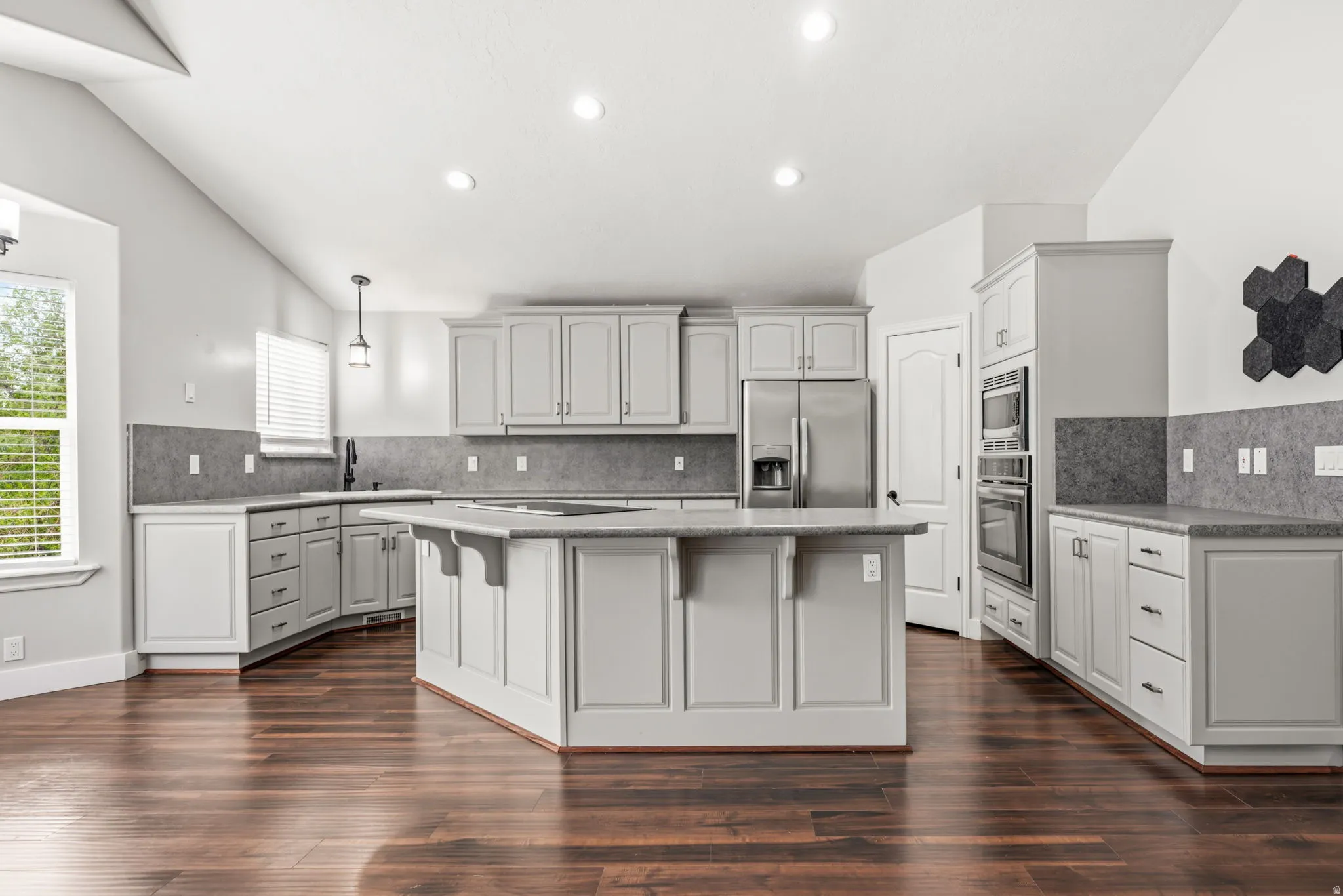 Spacious kitchen featuring an angled island, light gray cabinetry, stainless steel appliances, wood-finish flooring, and vaulted ceilings