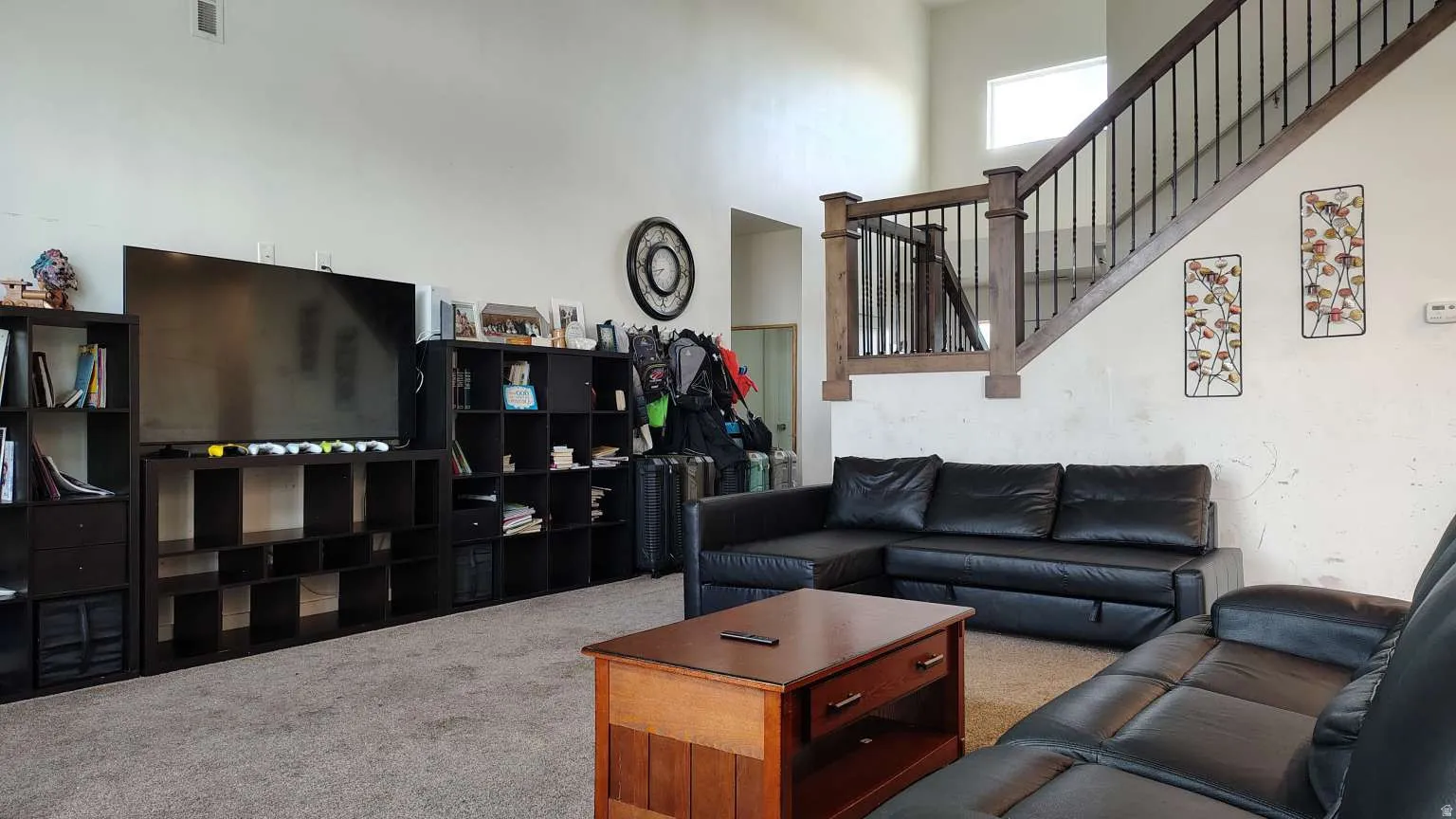 Carpeted living area with high ceilings, upper wall window, and a staircase featuring wood-finish handrails and black metal balusters