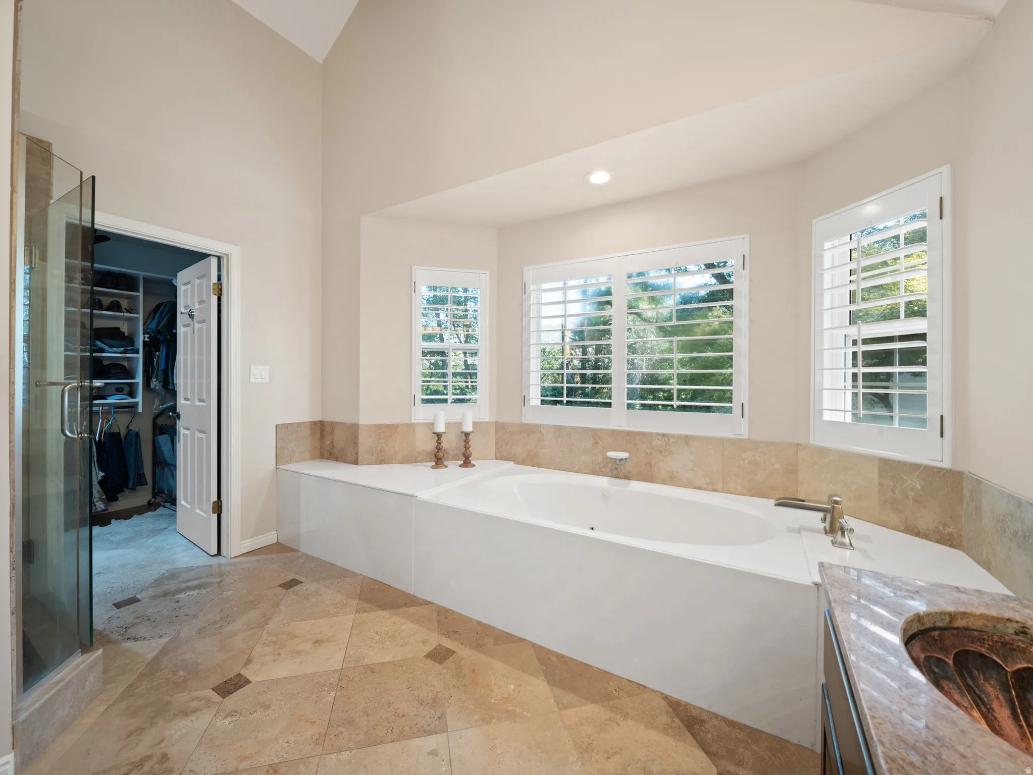 Deep soaking tub with surrounding stone tile, plantation shutters, vaulted ceiling, glass-enclosed shower, and stone tile flooring