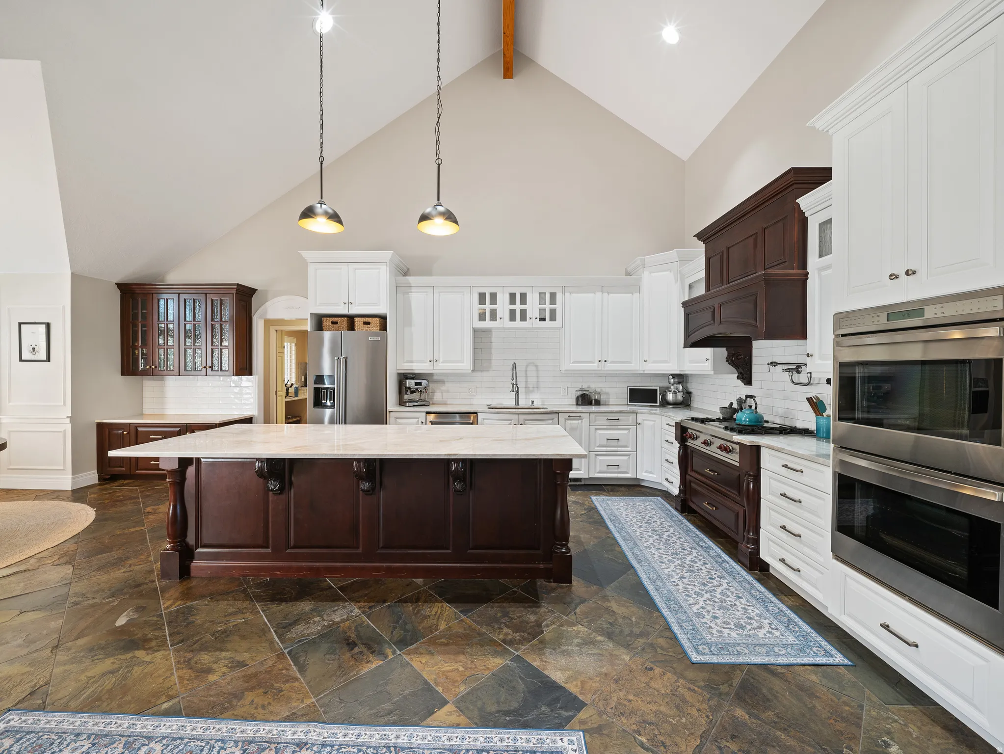Kitchen featuring a large central island with a light-toned countertop, vaulted ceiling with exposed wood beam, mixed white and dark wood cabinetry, subway tile backsplash, and slate-style flooring