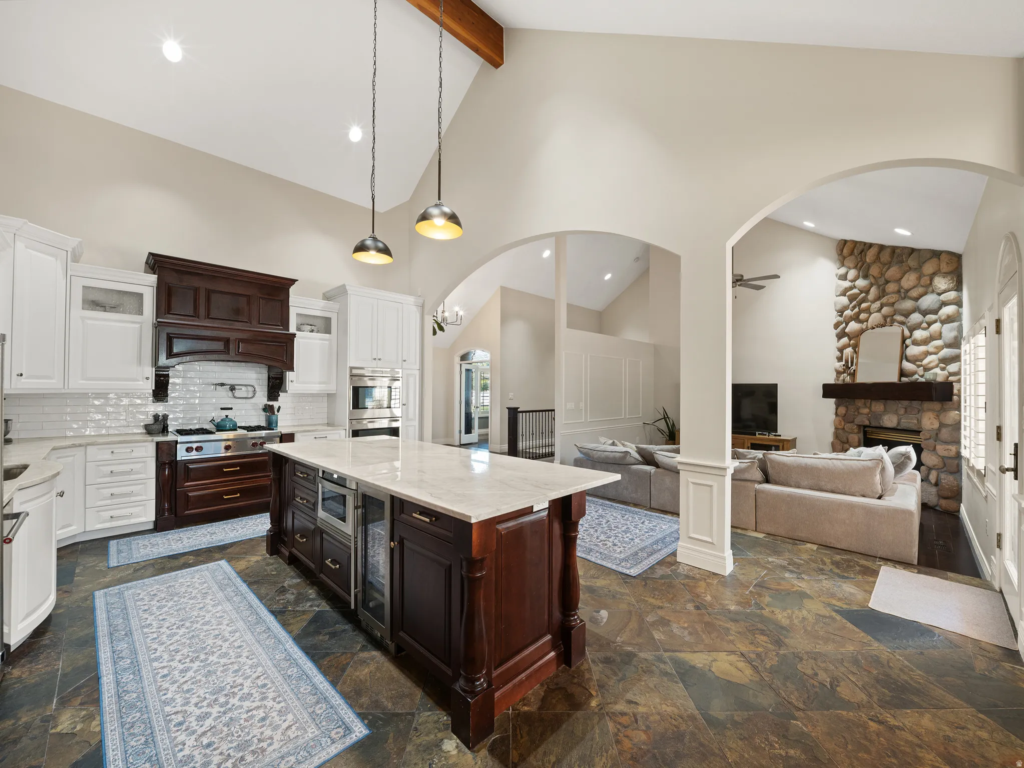 Gourmet kitchen featuring a large central island with integrated beverage refrigerator, vaulted ceilings with exposed wood beam, white cabinetry, dark wood stove hood, and slate tile flooring