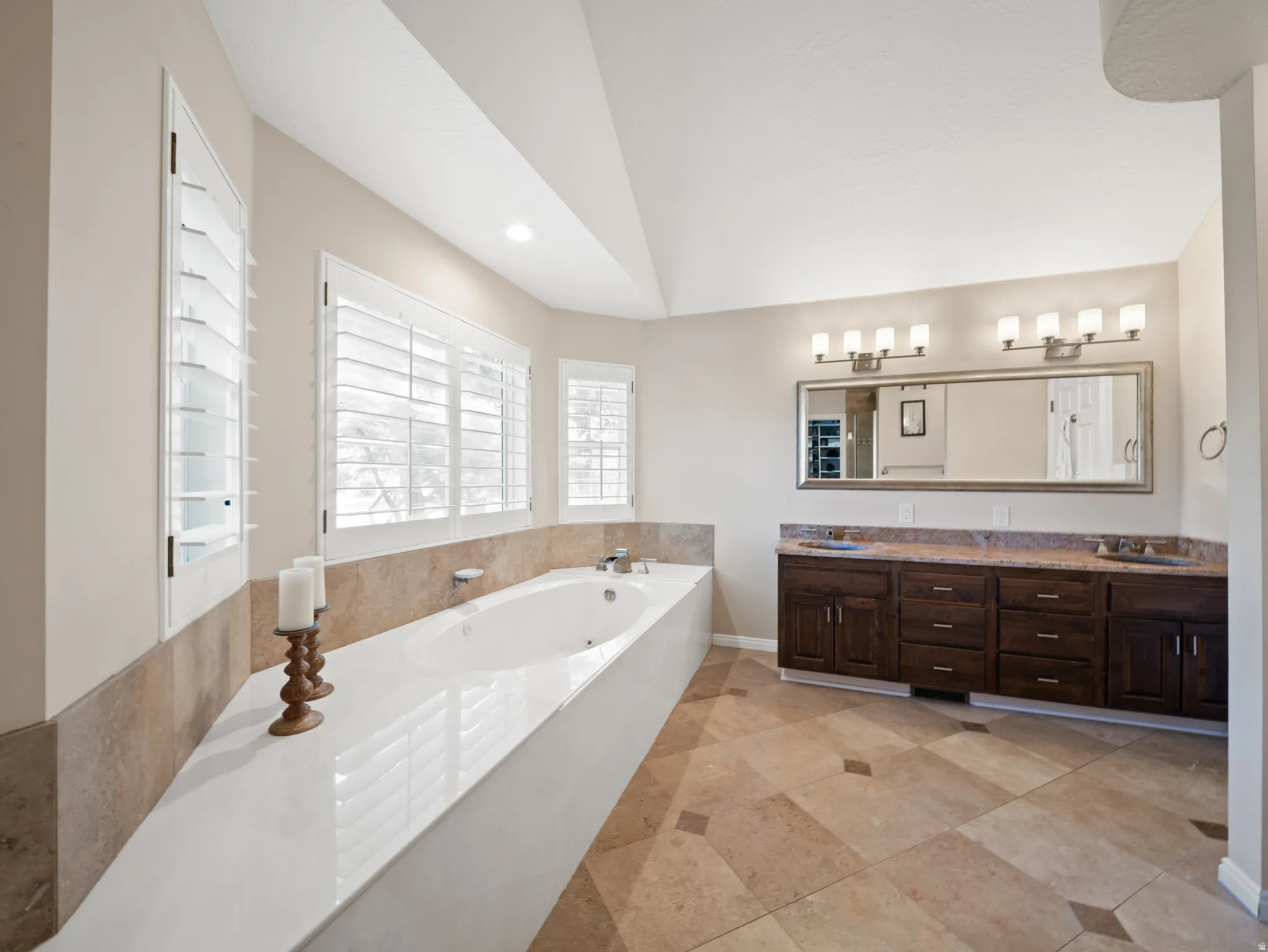 Soaking tub with a white surround and plantation shutters, complemented by a dual vanity with a stone countertop, dark wood-finish cabinetry, and a large mirror with sconce lighting