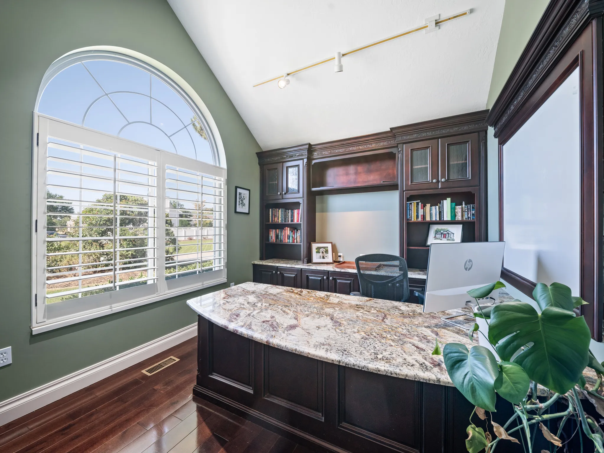 Arched window with plantation shutters, featuring a built-in desk with a stone countertop, custom cabinetry, and hardwood flooring