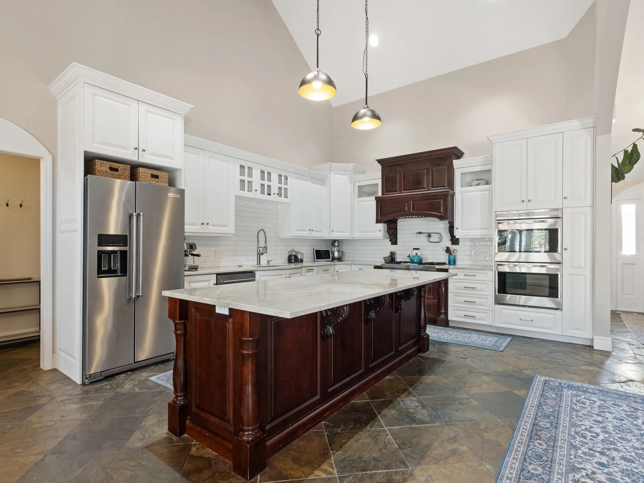 Expansive kitchen featuring a large central island with a light-toned countertop, white cabinetry, integrated stainless steel appliances, a decorative dark wood range hood, and slate flooring