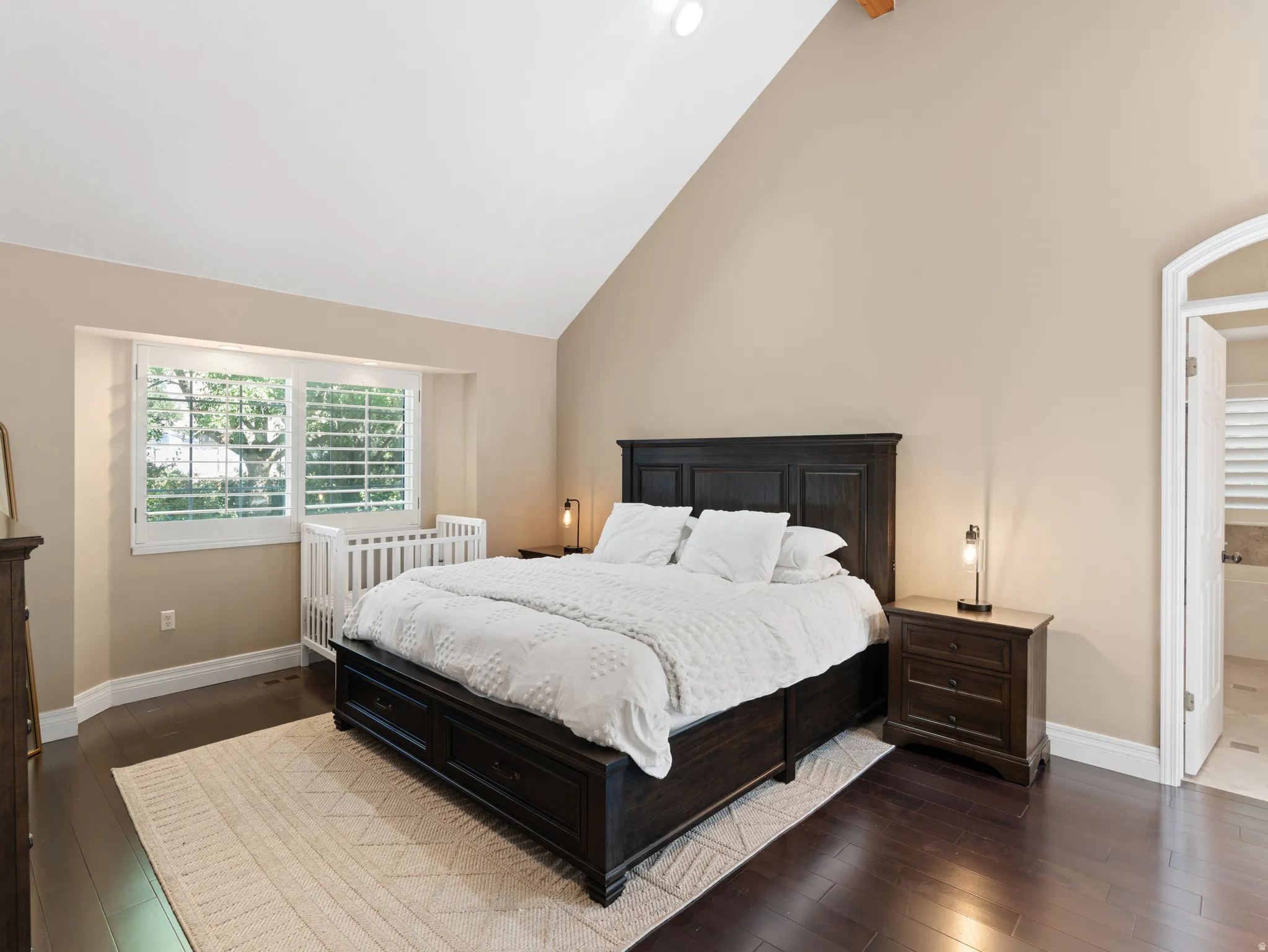 Spacious room featuring vaulted ceilings, dark wood-finish flooring, and a large window with white plantation shutters