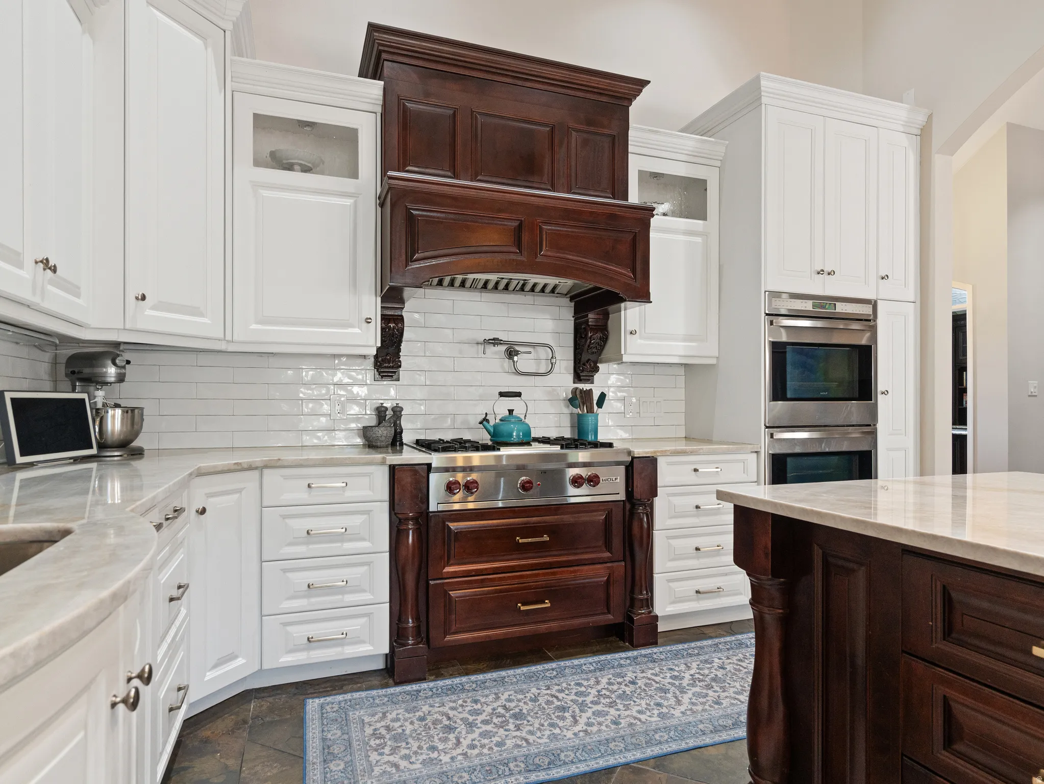 Gourmet kitchen featuring a wood-panel range hood, stainless steel gas range, white subway tile backsplash, light-toned countertops, and dual built-in stainless steel ovens