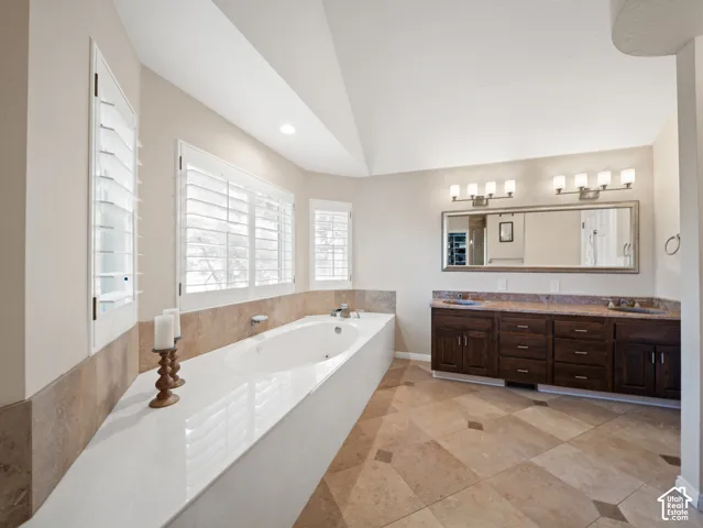 Soaking tub with a white surround and plantation shutters, complemented by a dual vanity with a stone countertop, dark wood-finish cabinetry, and a large mirror with sconce lighting