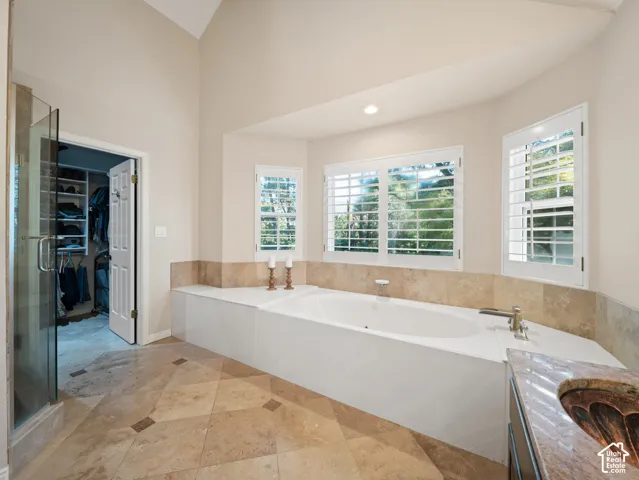 Deep soaking tub with surrounding stone tile, plantation shutters, vaulted ceiling, glass-enclosed shower, and stone tile flooring