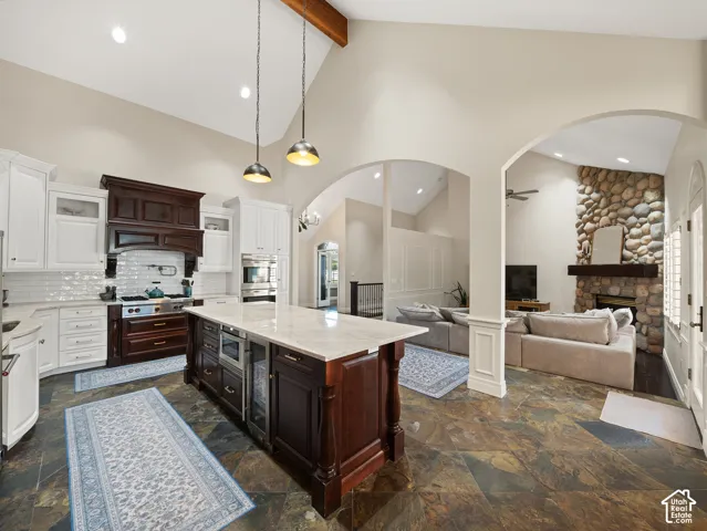 Gourmet kitchen featuring a large central island with integrated beverage refrigerator, vaulted ceilings with exposed wood beam, white cabinetry, dark wood stove hood, and slate tile flooring