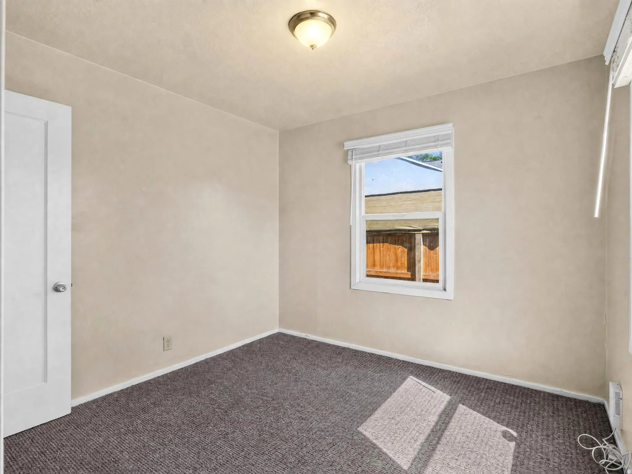 Neutral-toned interior room featuring a window with white blinds, grey patterned carpeting, and white baseboards