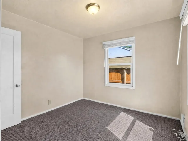 Neutral-toned interior room featuring a window with white blinds, grey patterned carpeting, and white baseboards