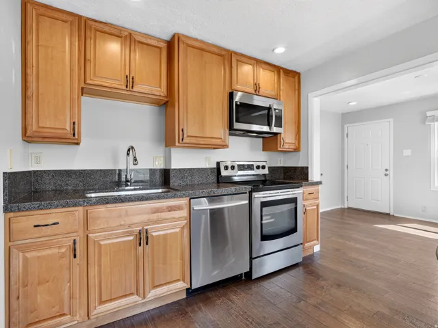 Kitchen featuring natural wood cabinetry, dark speckled countertops with backsplash, stainless steel appliances including a microwave, dishwasher, and electric range, and wood-finish flooring