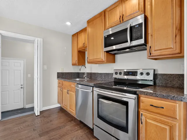 Kitchen featuring wood cabinetry, dark speckled countertops with a coordinating backsplash, stainless steel appliances, and dark wood-finish flooring