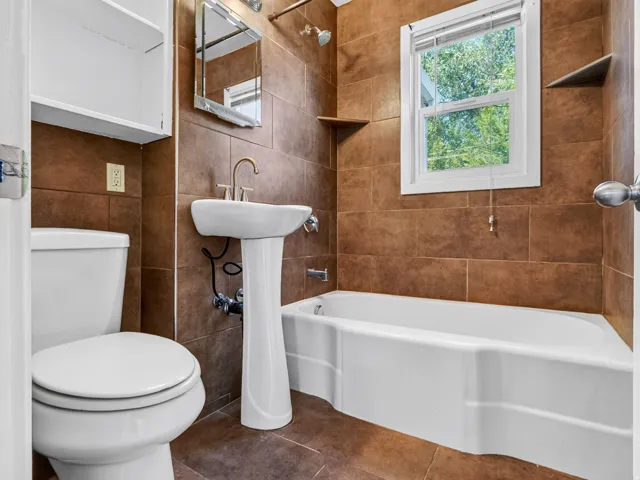 Bathroom featuring full-height brown tile walls and flooring, a white pedestal sink with brushed nickel faucet, a white toilet, and a white bathtub