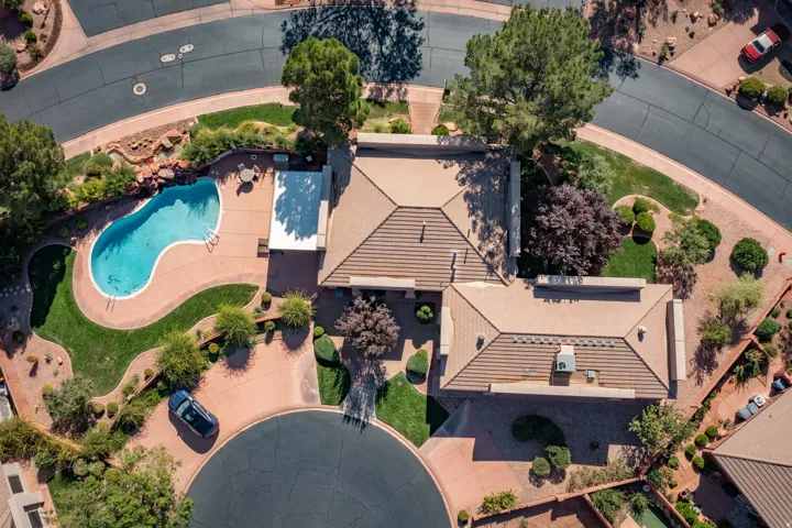 Aerial view of this residence showcasing a resort-style backyard retreat featuring a freeform swimming pool with integrated spa, dual covered pavilions with peaked rooflines, mature landscaping with specimen trees, circular hardscape elements, and meticul