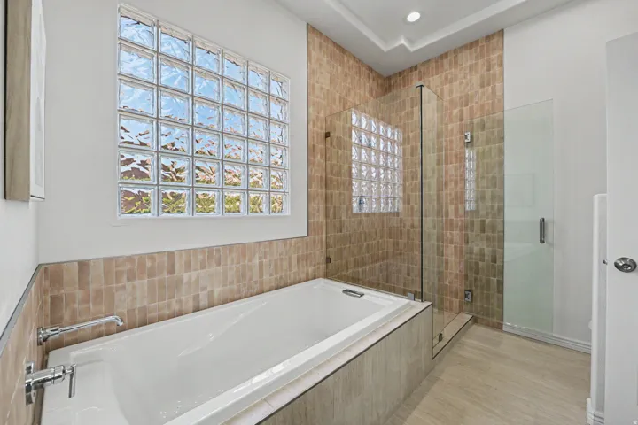 Bathroom featuring a built-in soaking tub with chrome fixtures, a glass block window, a walk-in shower with a glass enclosure, and stone-look wall and floor tiling