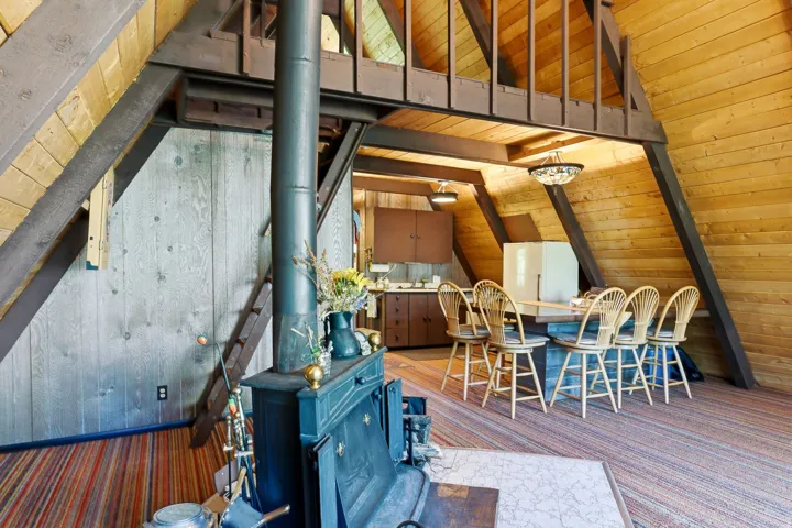 A-frame interior featuring natural wood plank walls, exposed dark wood beams, and a black wood-burning stove with a stovepipe