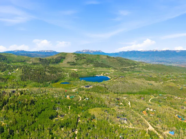 Expansive mountain landscape featuring dense green forests, multiple lakes, and distant snow-capped peaks