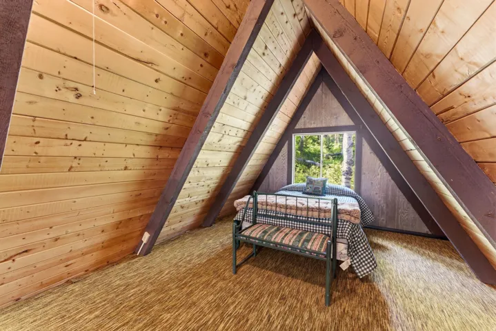 A-frame interior featuring natural wood plank walls, exposed timber beams, a fixed window with a wooded backdrop, and textured carpeting
