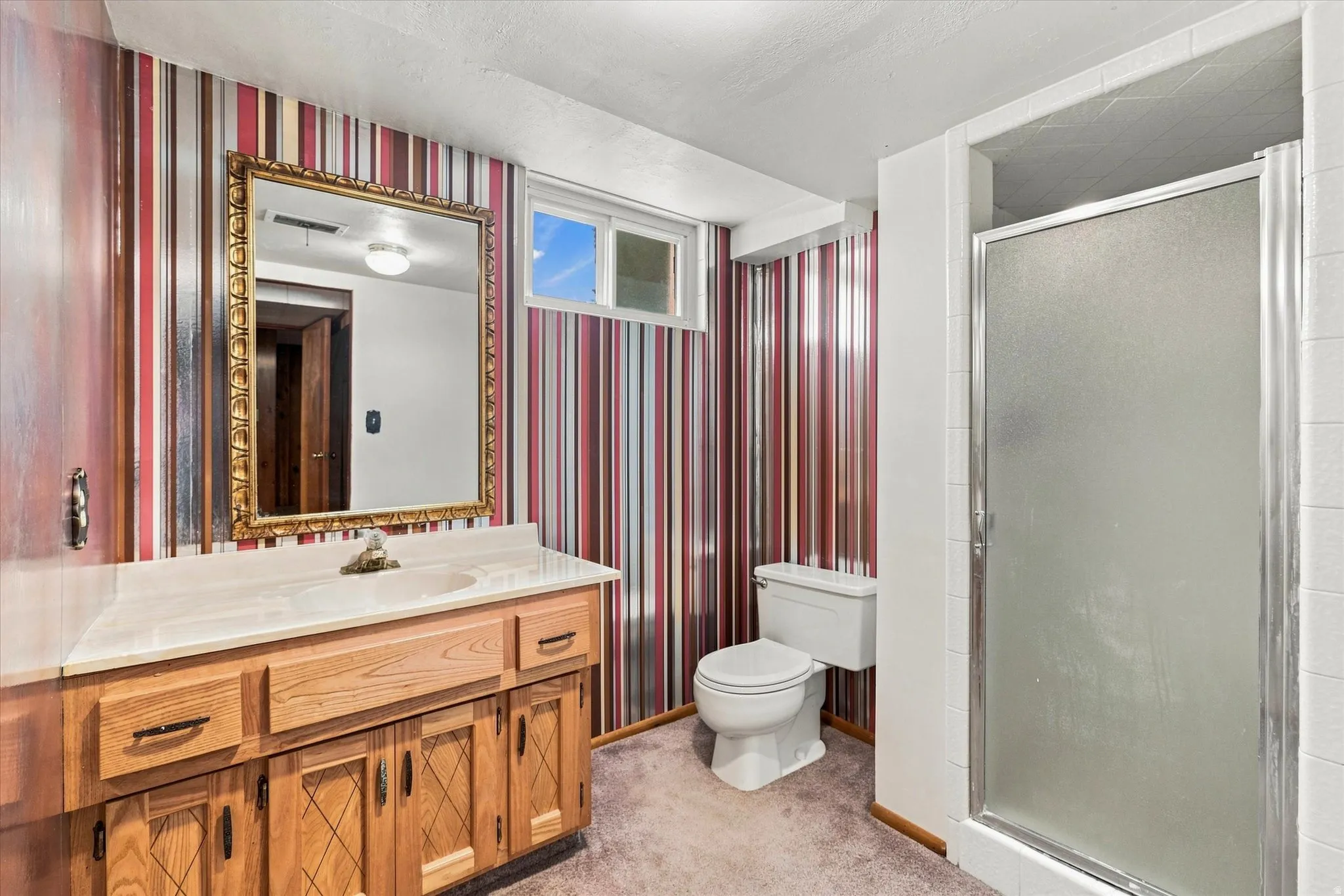 Bathroom featuring a wood vanity with a light-toned countertop and dual sinks, a decorative gold-framed mirror, a frosted glass shower enclosure, and a high horizontal window