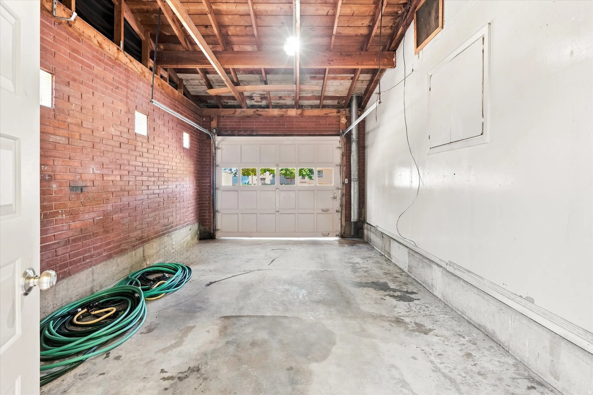 Spacious two-car garage featuring a white overhead door with windows, exposed wood beam ceiling, brick accent wall, and concrete flooring