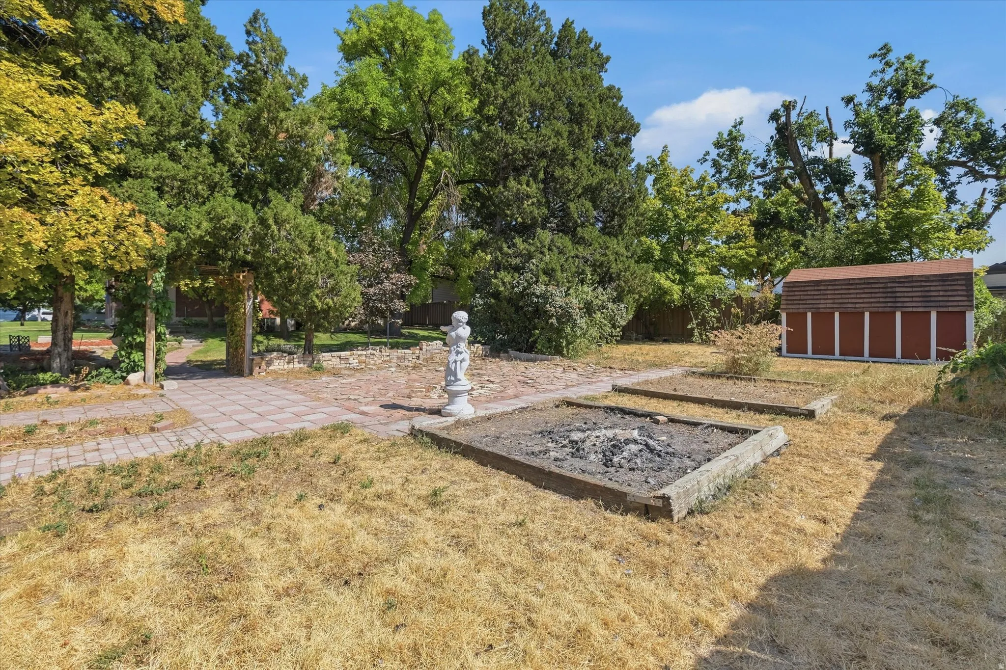Expansive outdoor space featuring a paved walkway, mature trees, a garden shed, raised planter beds, and an integrated fire pit