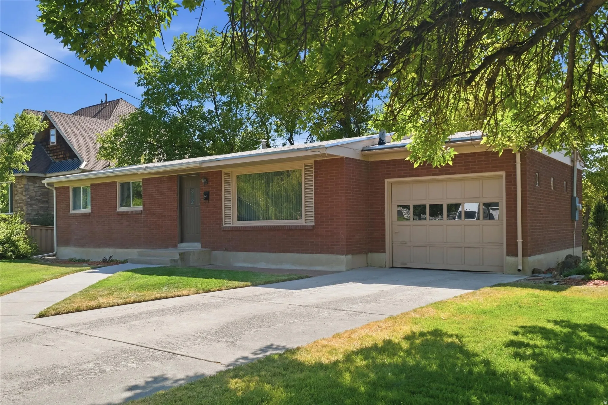Brick exterior home featuring a concrete driveway, attached garage with panel door and window inserts, front entry with covered porch, and mature trees framing the property