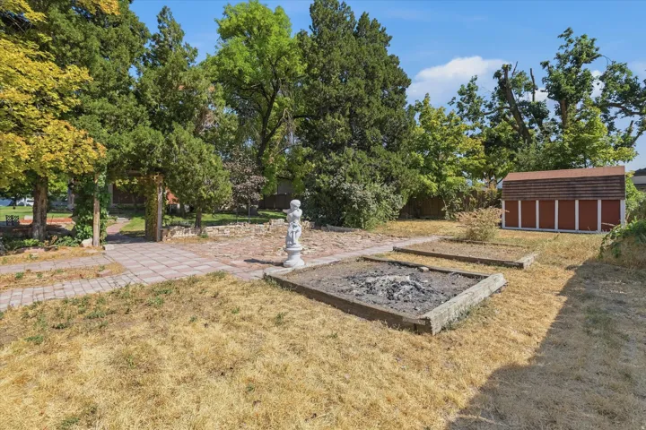 Expansive outdoor space featuring a paved walkway, mature trees, a garden shed, raised planter beds, and an integrated fire pit