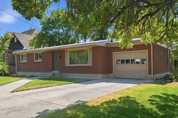Brick exterior home featuring a concrete driveway, attached garage with panel door and window inserts, front entry with covered porch, and mature trees framing the property