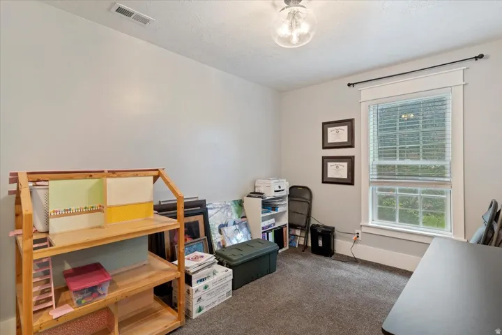 Versatile bedroom or multi-purpose room featuring wood-frame shelving with storage capacity, carpeted flooring, large window with plantation shutters and natural light, ceiling-mounted lighting fixture, and white painted walls
