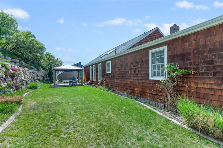 Charming cedar-shingled home with a spacious backyard featuring manicured lawn, mature landscaping with flowering shrubs, and a covered gazebo structure
