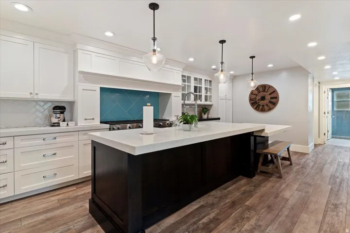 Kitchen featuring a large central island with a light-toned countertop, white perimeter cabinetry, a vibrant blue tile backsplash, stainless steel range, and wood-finish flooring