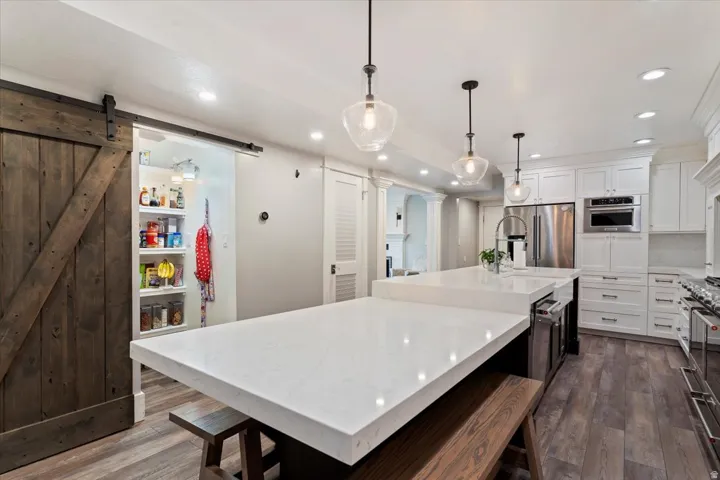 Gourmet kitchen featuring a large waterfall-edge quartz island with seating, white cabinetry, stainless steel appliances, pendant lighting, wood-finish flooring, and a sliding barn door pantry with open shelving