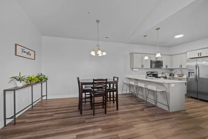 Open-concept kitchen and dining area featuring stainless steel appliances, white cabinetry with hardware accents, quartz countertops, a central island with seating, pendant lighting, wood-finish flooring throughout, and a spacious layout with high ceiling
