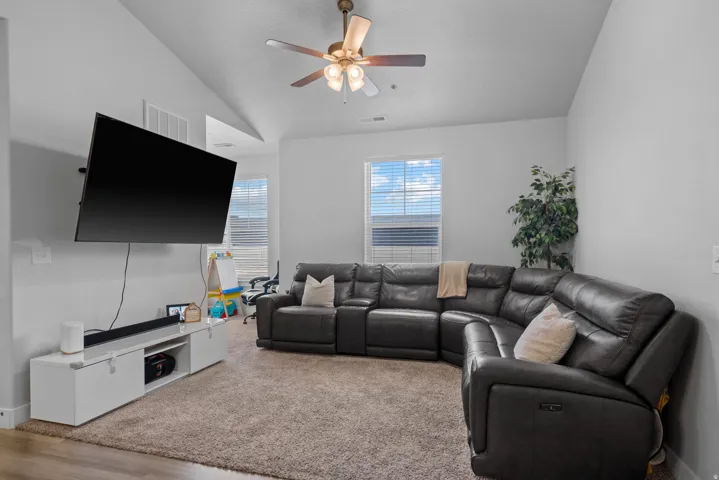 Spacious entertainment room featuring a sectional leather seating arrangement, ceiling fan with integrated lighting, wall-mounted flat-screen television, engineered wood-finish flooring, and abundant natural light from multiple windows