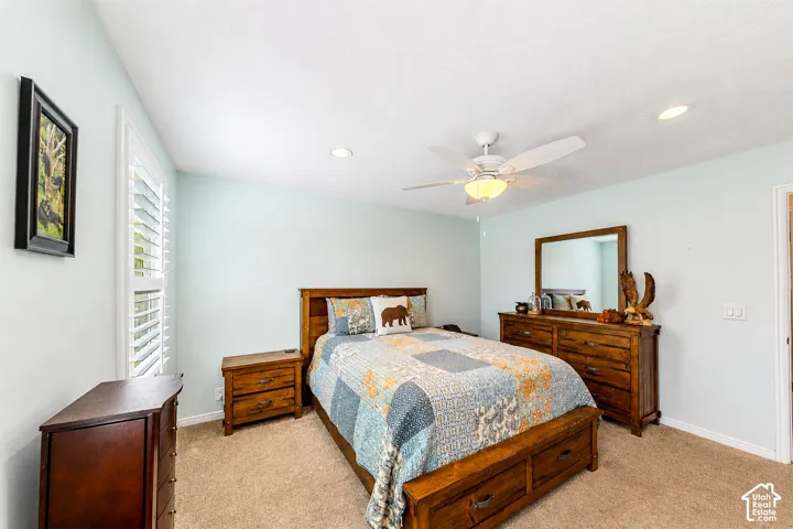 Spacious bedroom featuring warm wood-finish furnishings, neutral carpeting, and soft ambient lighting from a ceiling fan with integrated fixture
