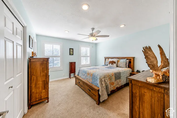 Carpeted bedroom featuring light blue walls, two windows with white plantation shutters, a ceiling fan with integrated lighting, and recessed ceiling lights